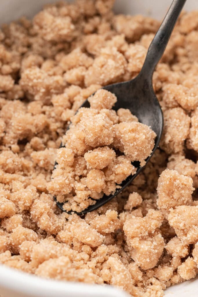 A spoon scooping crumbly brown sugar and flour streusel topping from a bowl.
