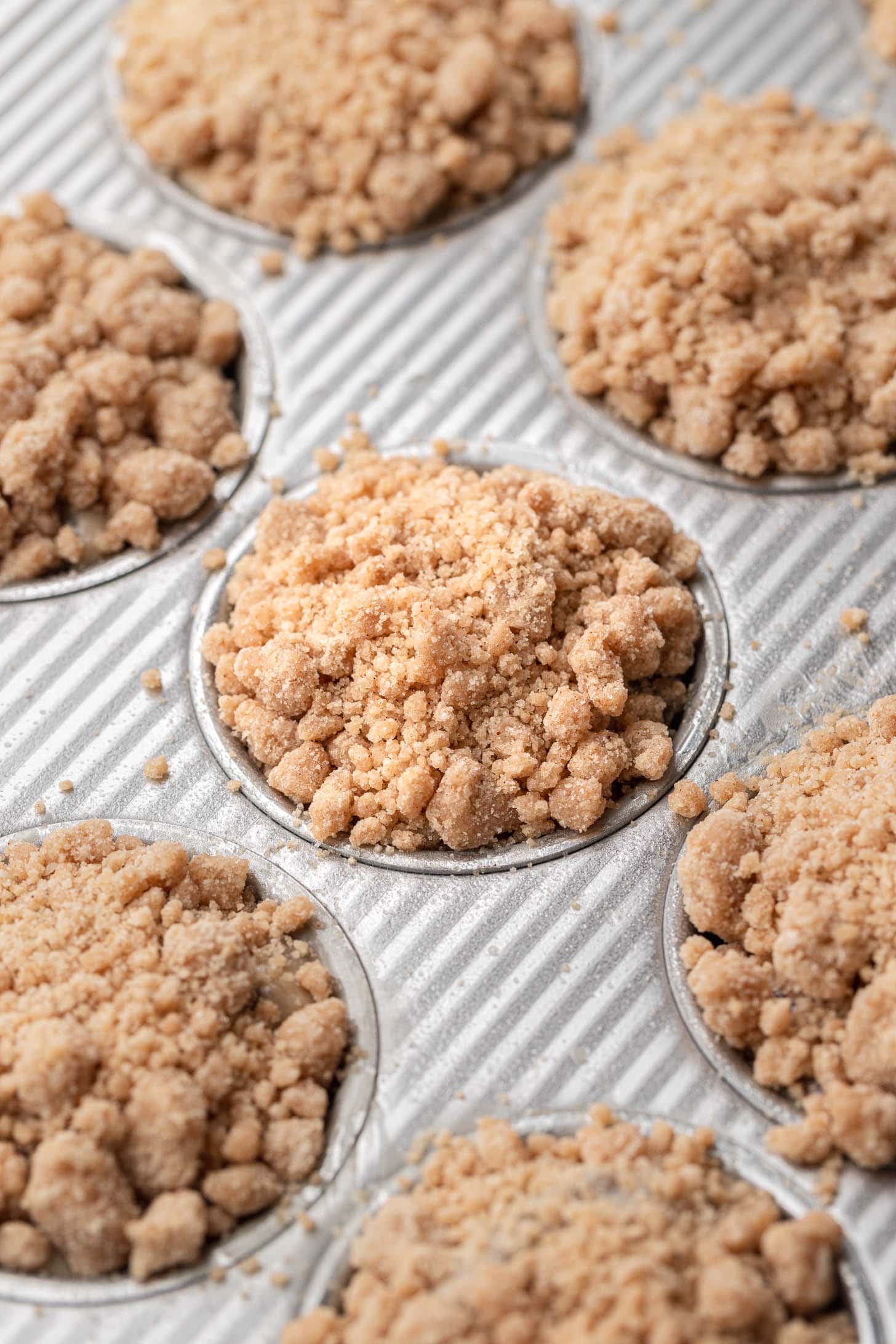 Unbaked crumb-topped muffins in a metal muffin tin, ready to go into the oven.