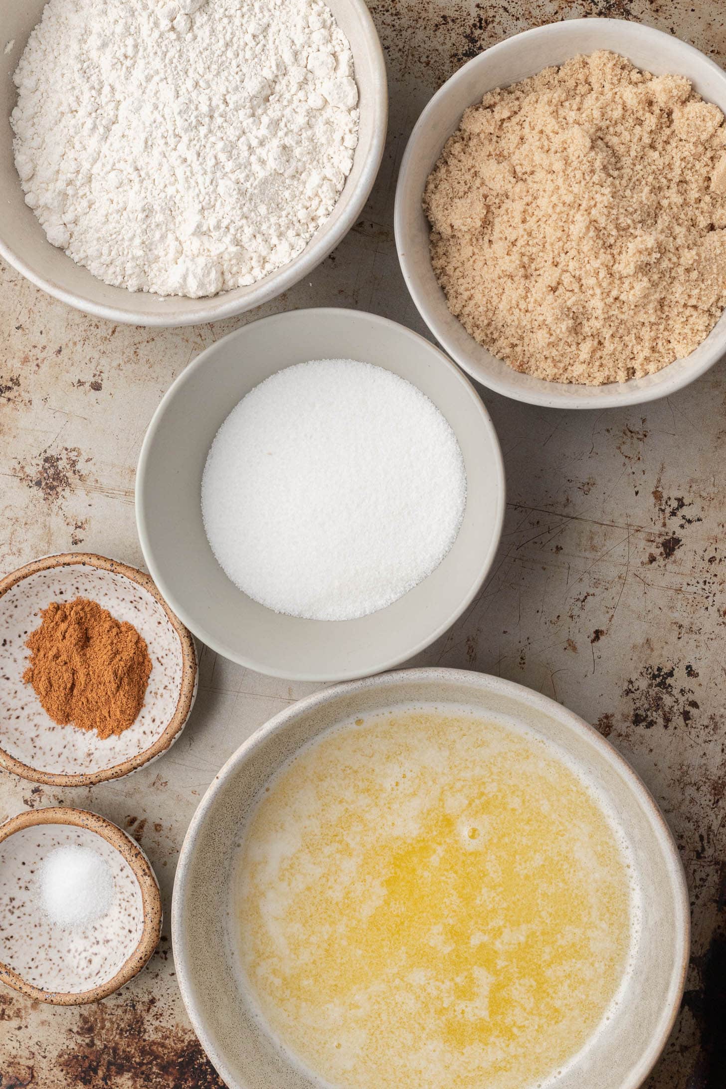 Bowls with flour, brown sugar, white sugar, melted butter, cinnamon, and salt on a rustic surface.