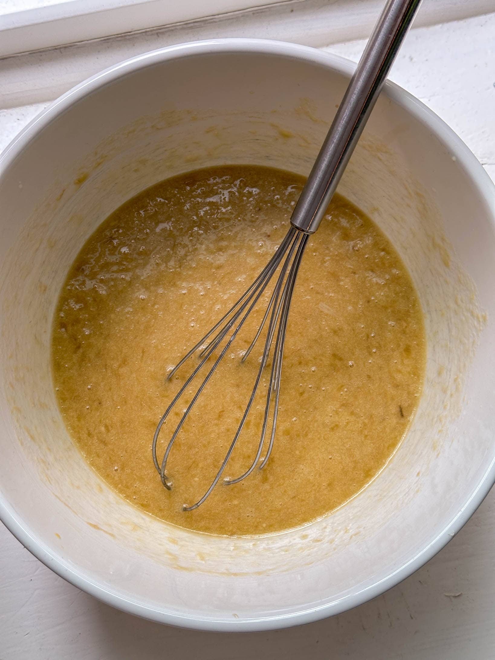 A metal whisk in a bowl of yellowish batter on a white surface.