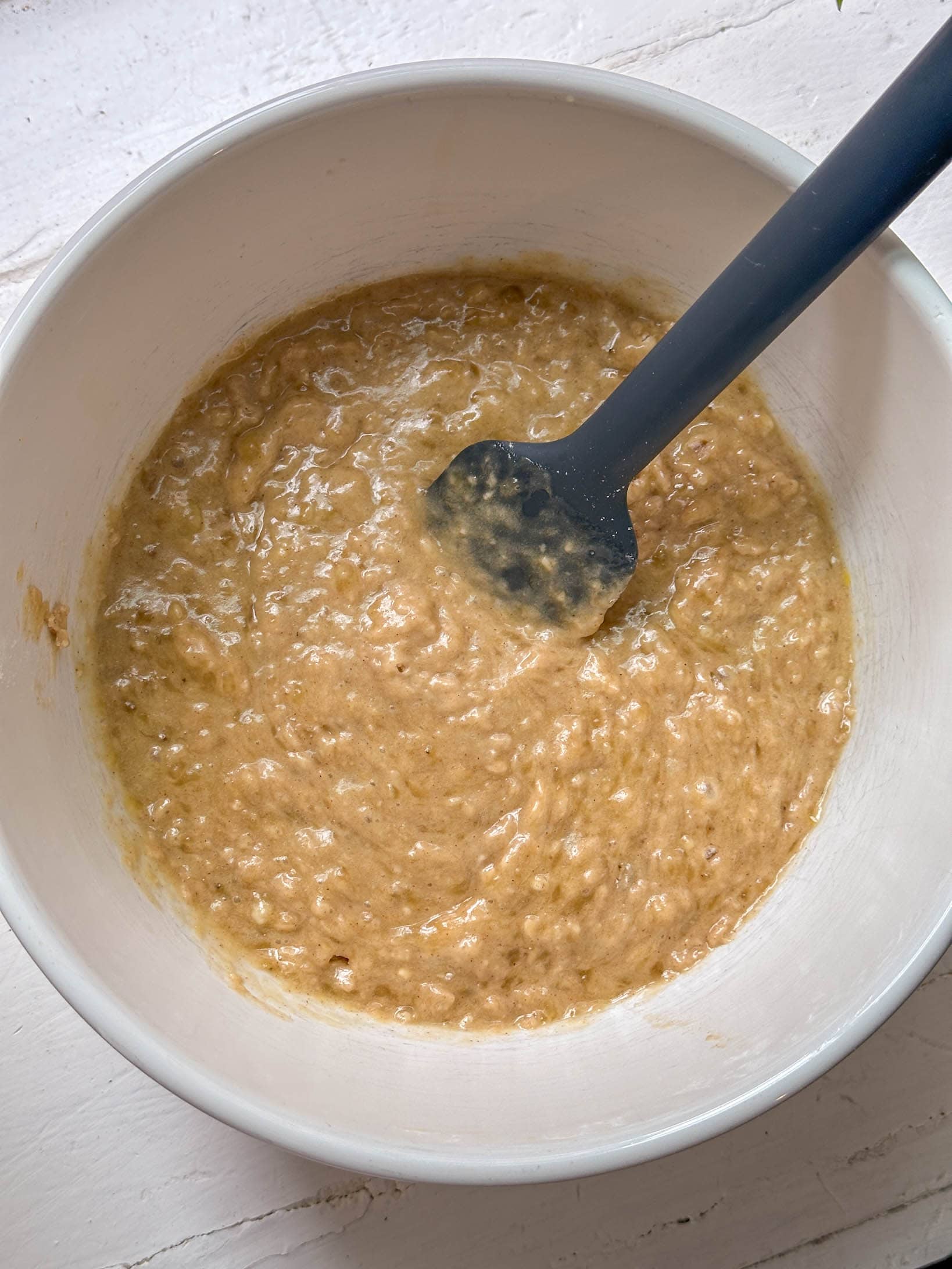 A mixing bowl with lumpy banana muffin batter and a black spatula on a white surface.
