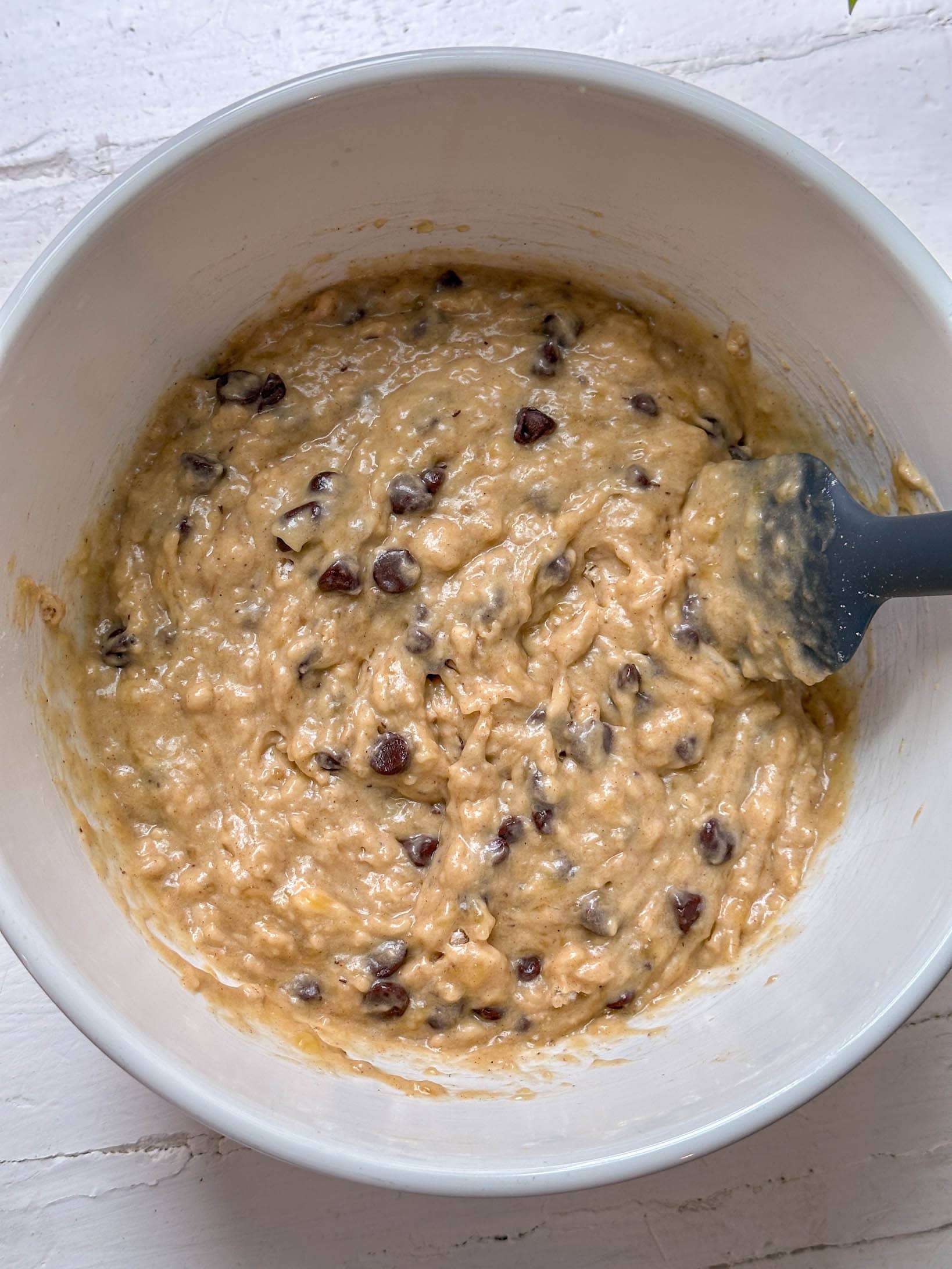 Banana bread batter with chocolate chips in a white bowl, stirred with a black spatula on a white surface.