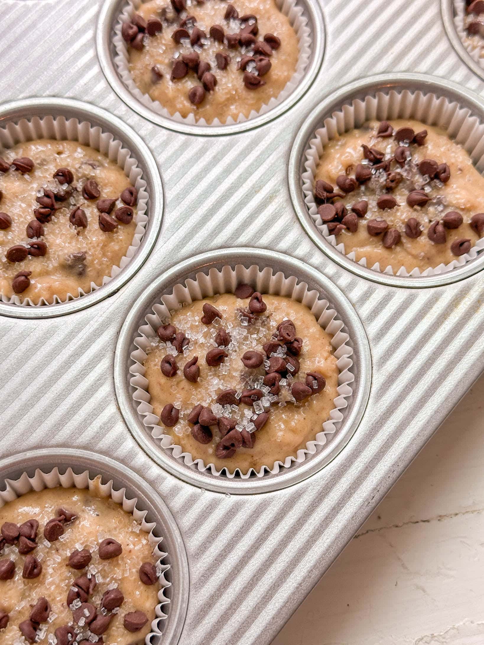 Muffin batter in cupcake liners topped with mini chocolate chips and sugar, ready to bake in a muffin tin.