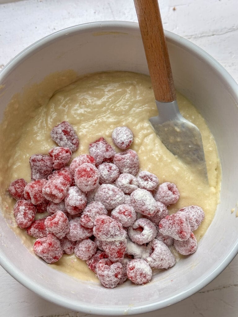 A bowl of muffin batter with flour-coated raspberries and a spatula on a white surface.