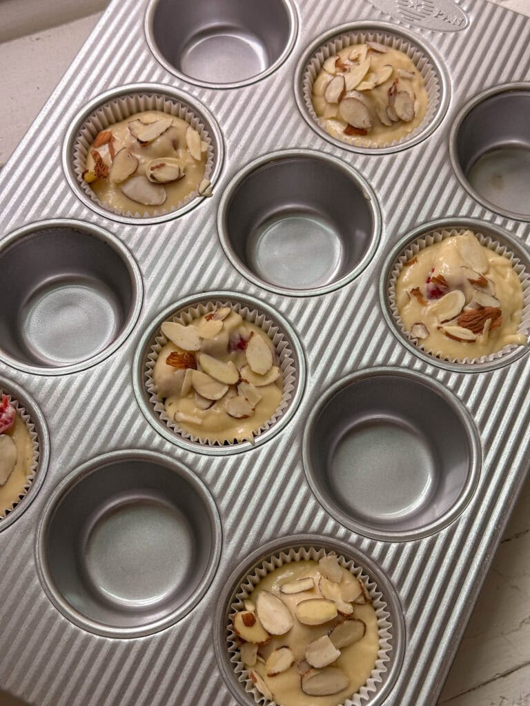 A muffin tin with a few unbaked, almond-topped muffins in paper liners; the rest of the cups are empty.