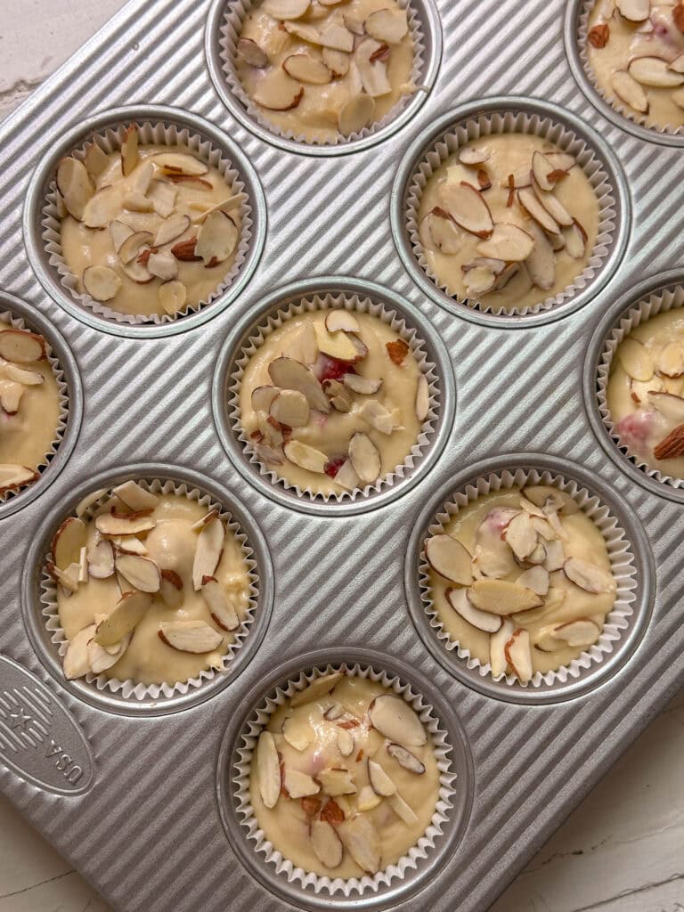 Muffin tin filled with unbaked muffin batter topped with sliced almonds, ready to be baked.