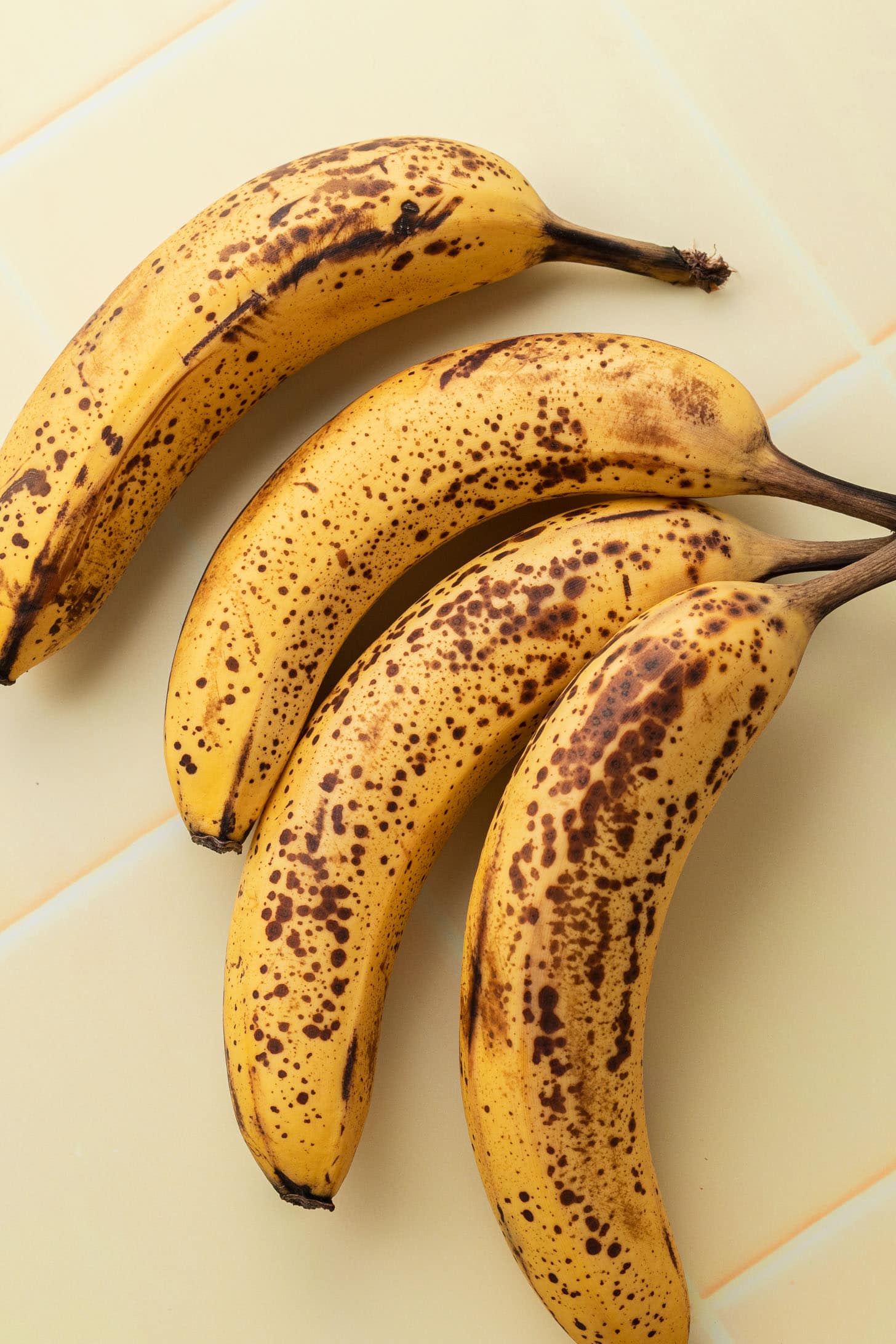 Four ripe bananas with brown spots on a light yellow tiled surface.
