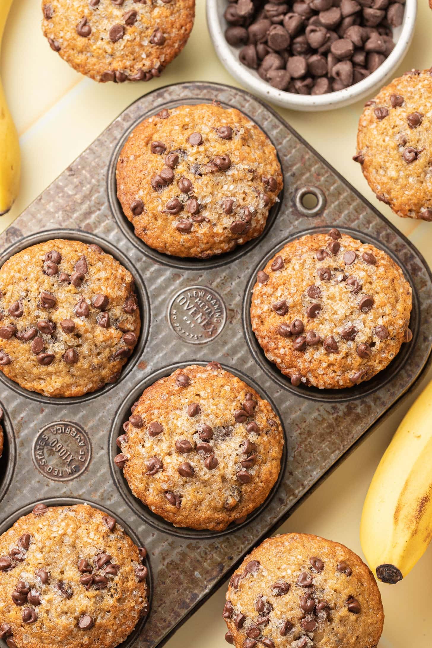 Banana chocolate chip muffins in a muffin tin, surrounded by fresh bananas and a bowl of chocolate chips.