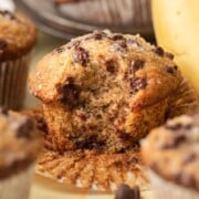 A banana chocolate chip muffin with a bite taken out, surrounded by more muffins and a banana.