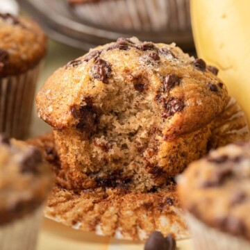 A banana chocolate chip muffin with a bite taken out, surrounded by more muffins and a banana.