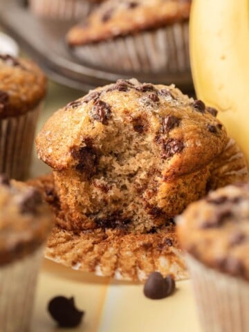 A banana chocolate chip muffin with a bite taken out, surrounded by more muffins and a banana.