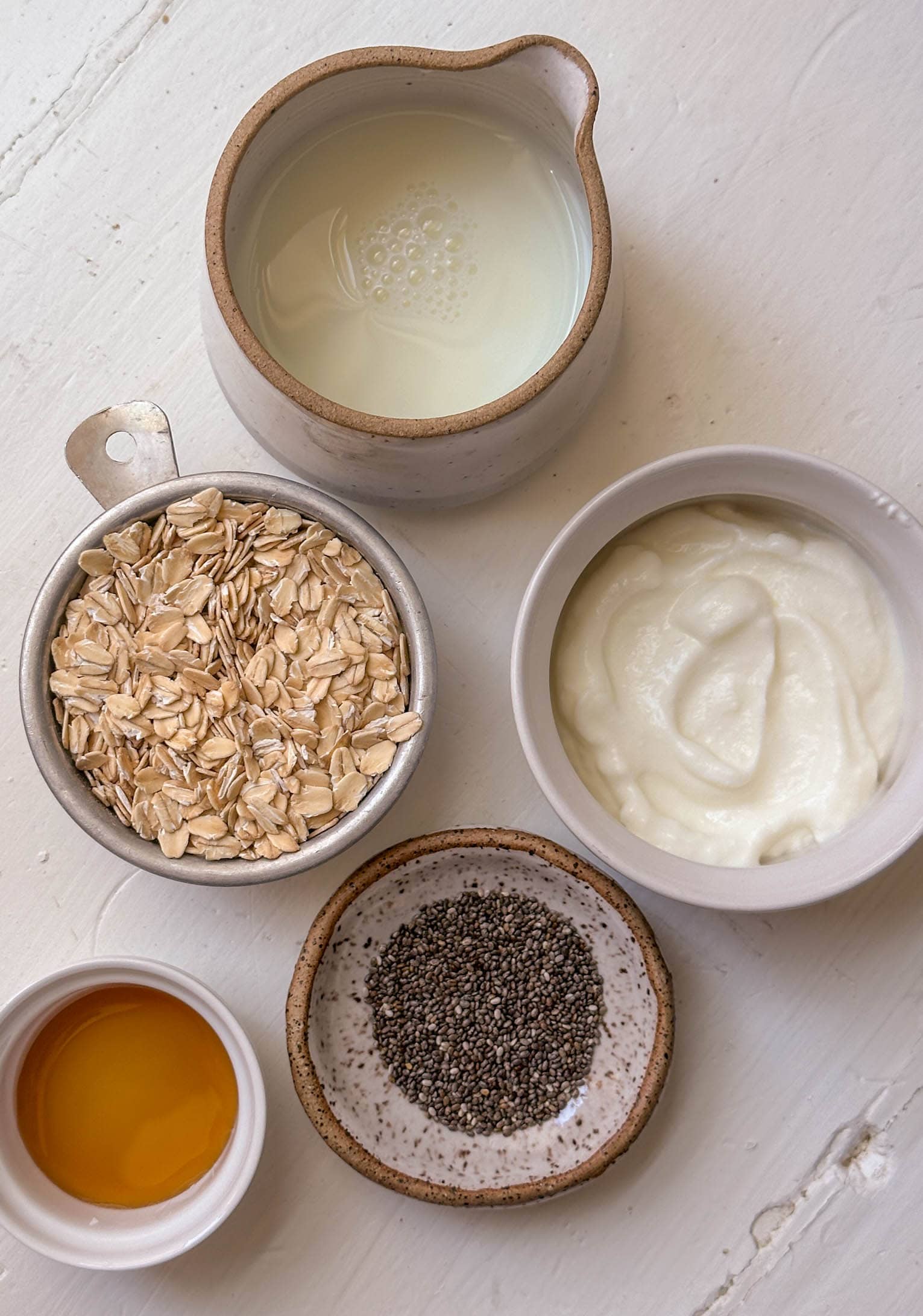 Five bowls with milk, yogurt, rolled oats, chia seeds, and maple syrup arranged on a white surface.