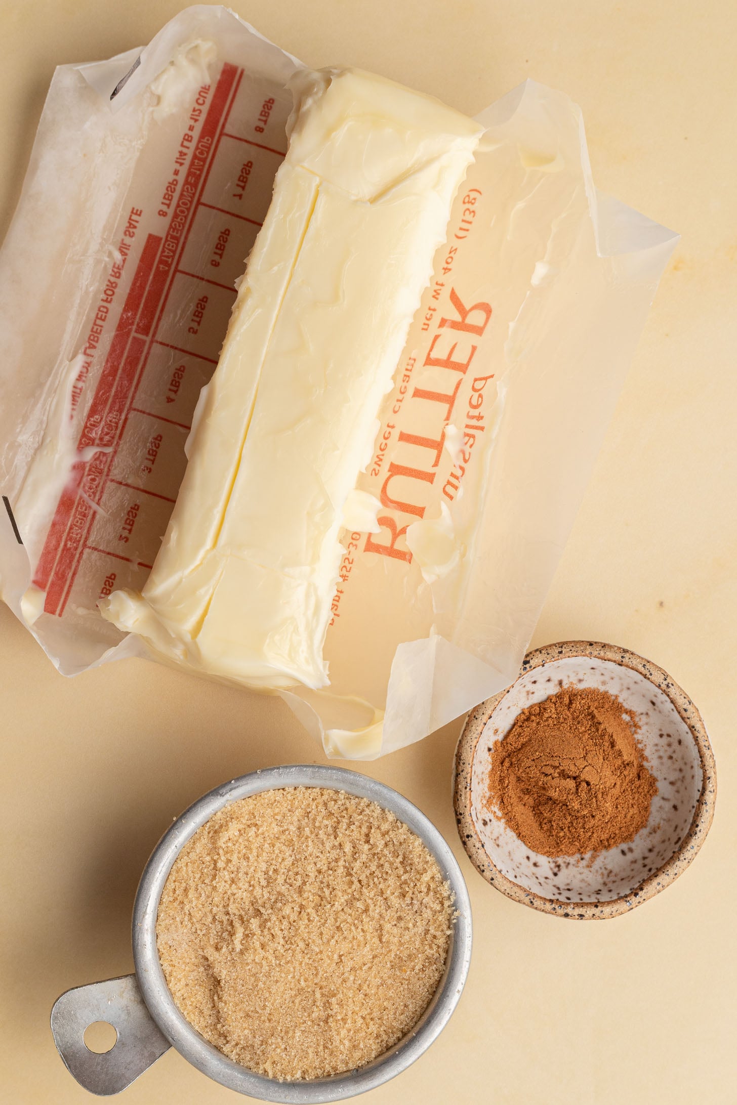 Unwrapped butter, a small bowl of ground cinnamon, and a measuring cup of brown sugar on a beige surface.