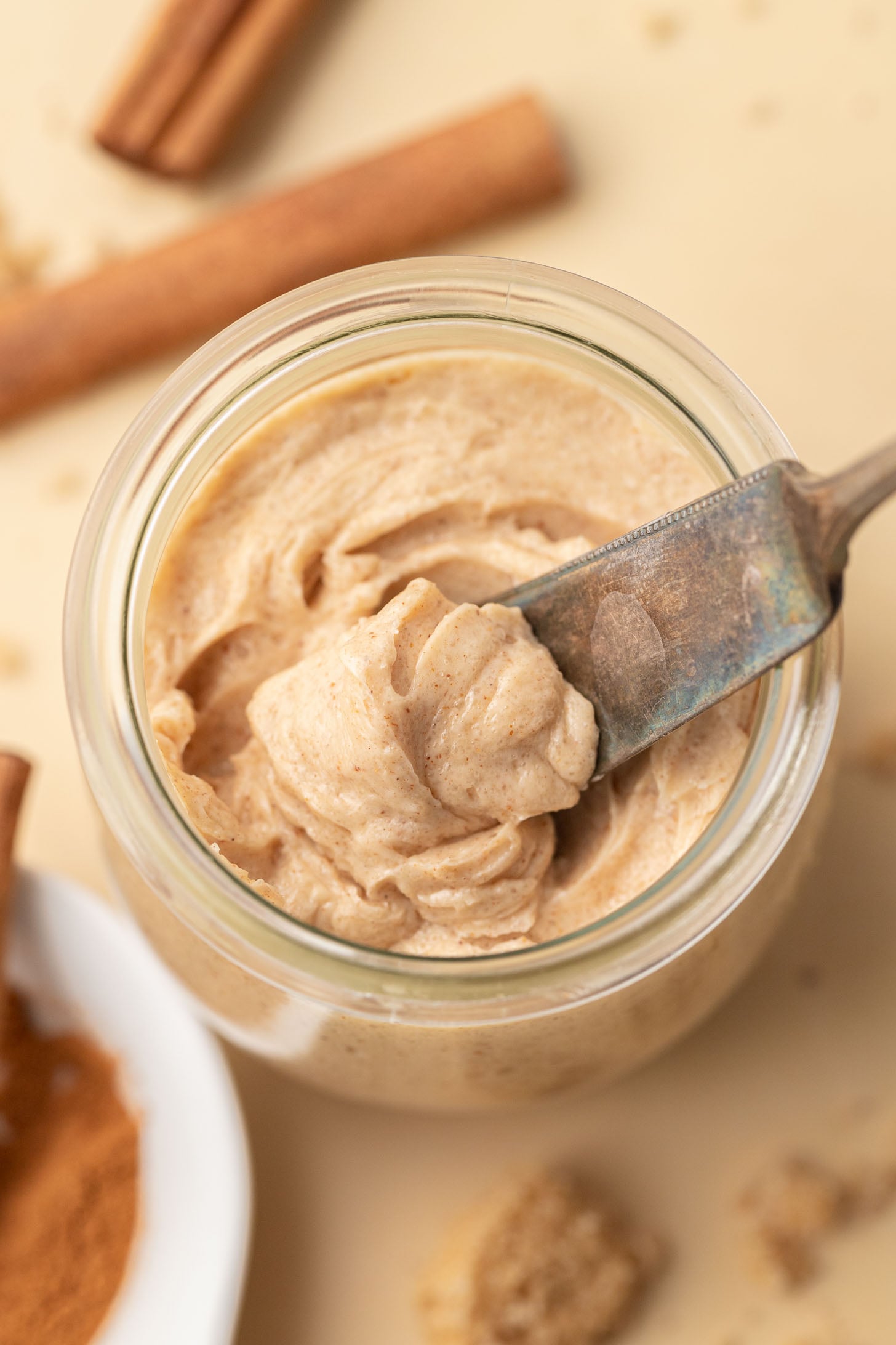 A jar of creamy cinnamon butter with a knife, surrounded by cinnamon sticks and brown sugar.