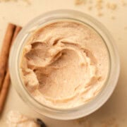 A jar of whipped cinnamon butter surrounded by cinnamon sticks and brown sugar on a light surface.