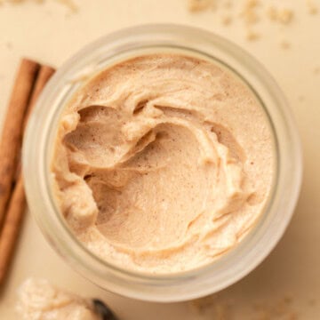 A jar of whipped cinnamon butter surrounded by cinnamon sticks and brown sugar on a light surface.