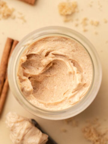 A jar of whipped cinnamon butter surrounded by cinnamon sticks and brown sugar on a light surface.