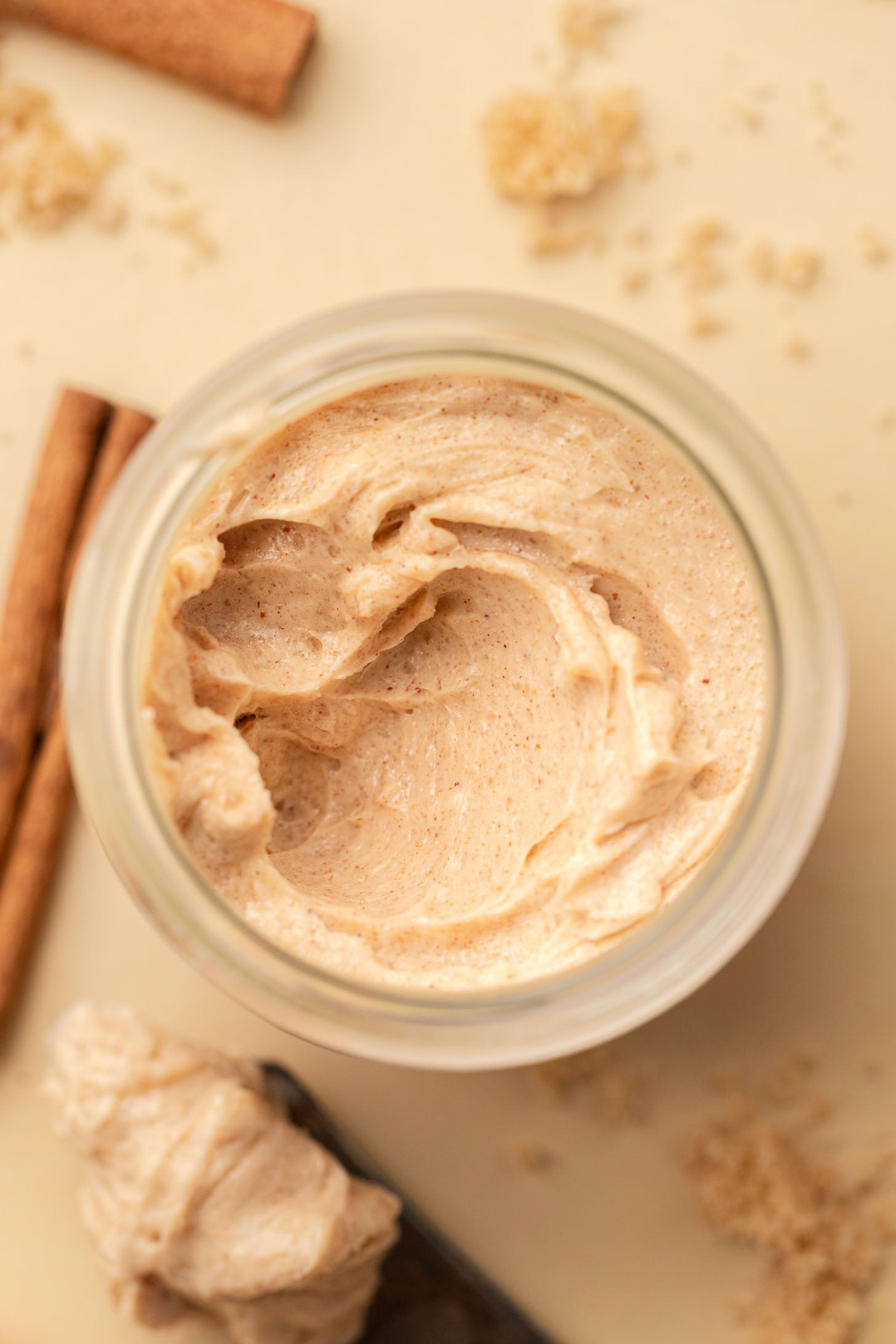 A jar of whipped cinnamon butter surrounded by cinnamon sticks and brown sugar on a light surface.