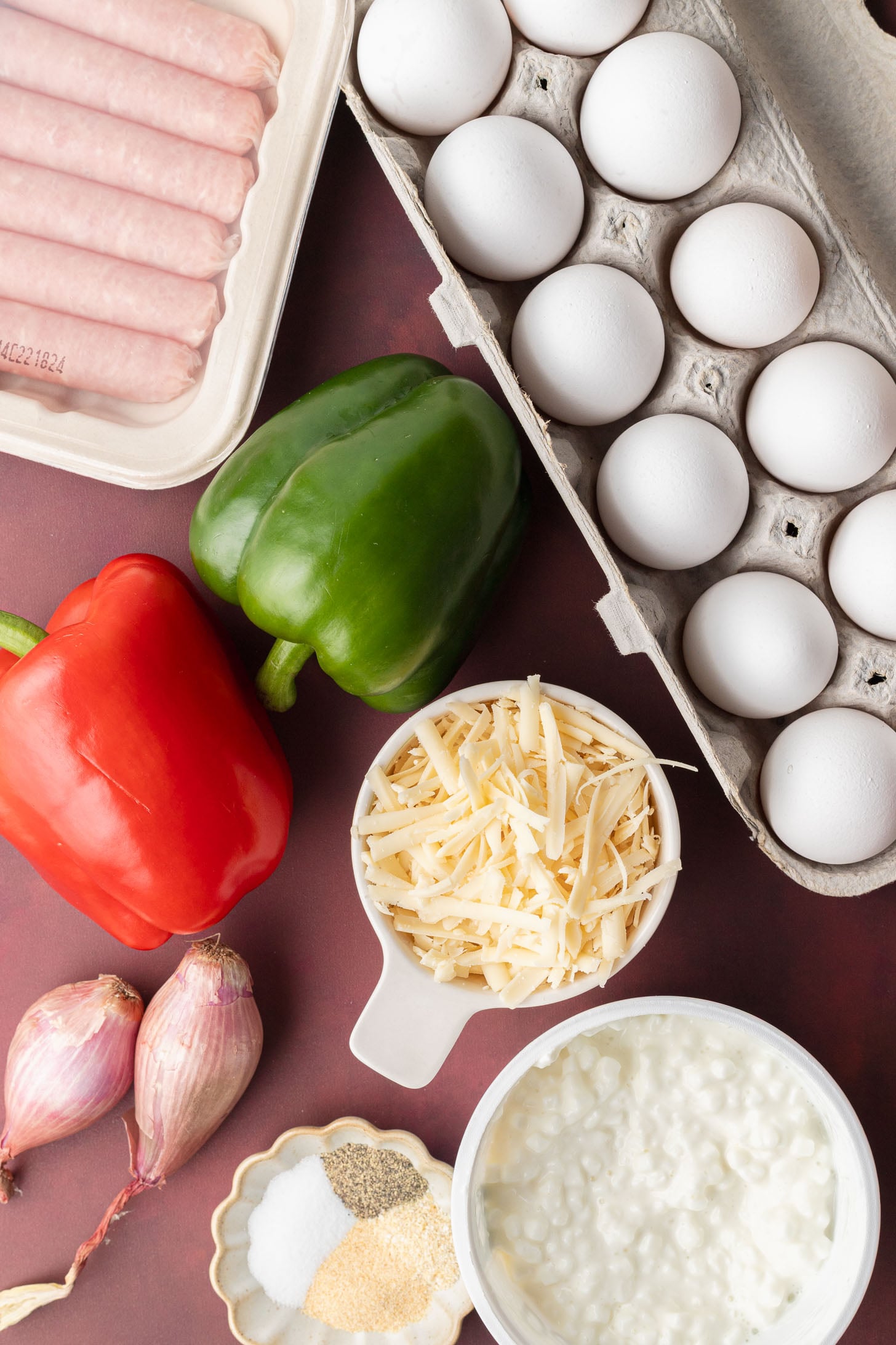 Eggs, raw sausages, red and green bell peppers, shredded cheese, shallots, cottage cheese, and spices on a table.