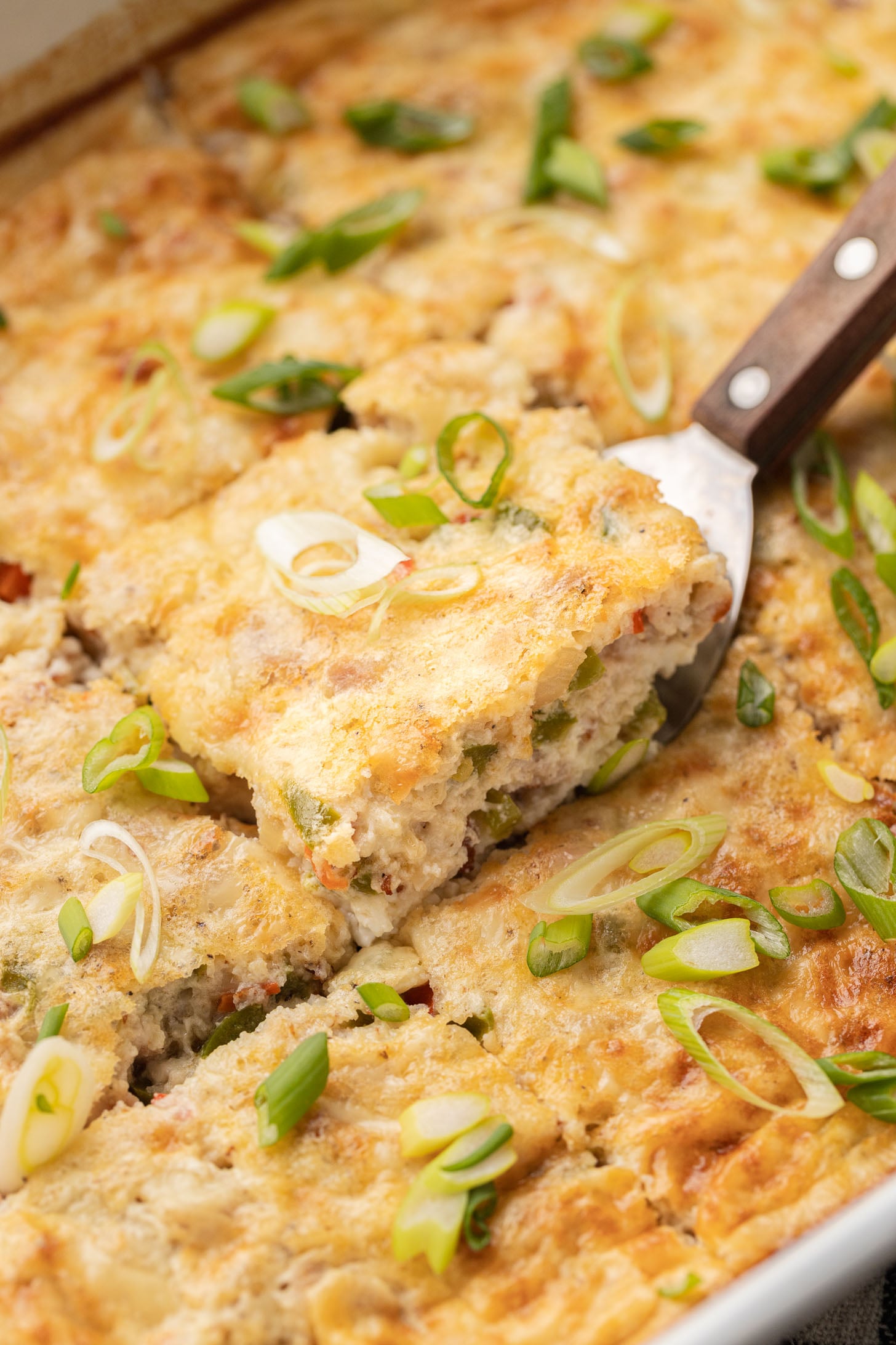 A spatula lifts a slice of cheesy casserole topped with chopped green onions from a baking dish.