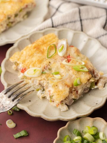 A slice of baked casserole topped with sliced green onions on a plate, with a fork and more onions nearby.