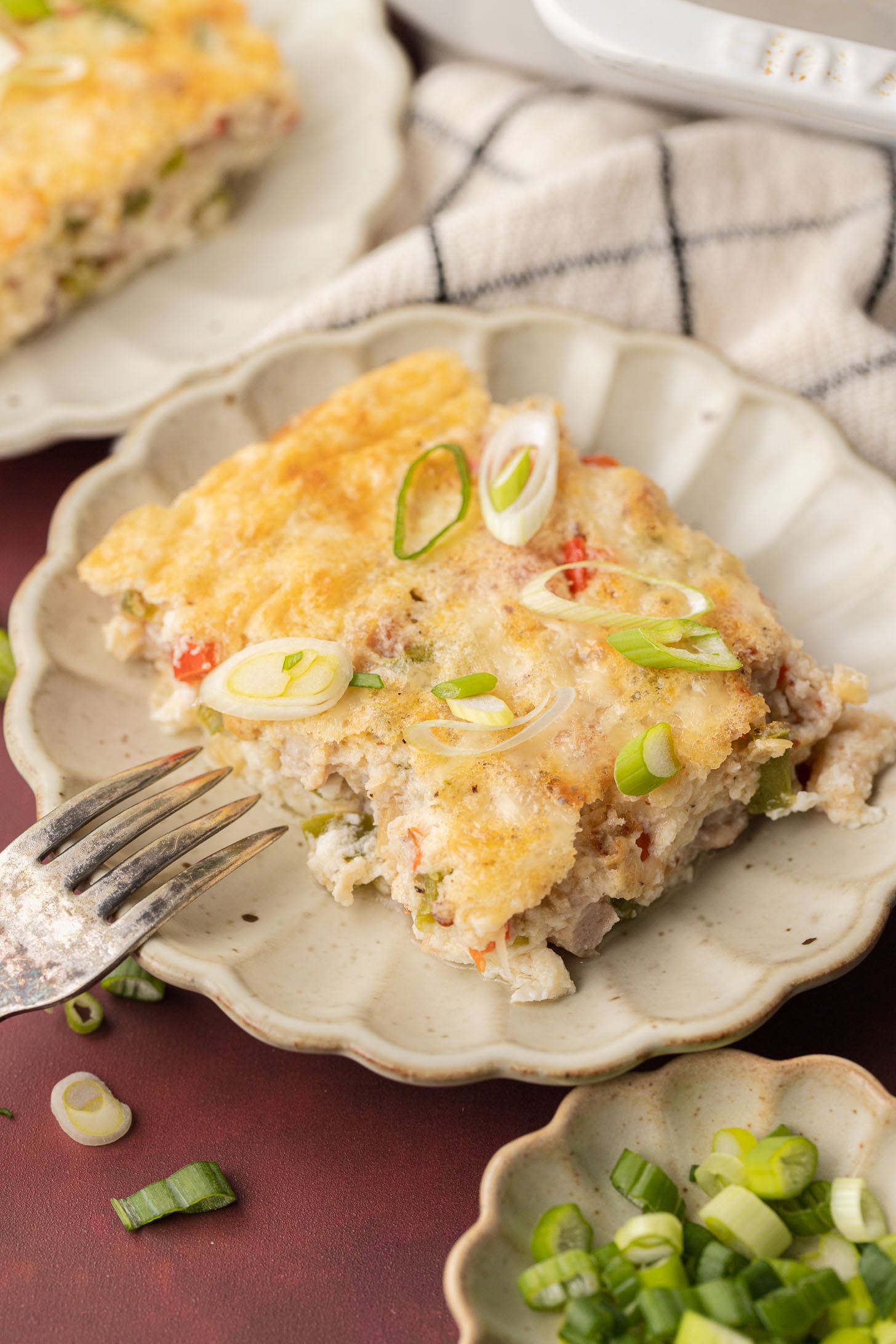 A slice of baked casserole topped with sliced green onions on a plate, with a fork and more onions nearby.
