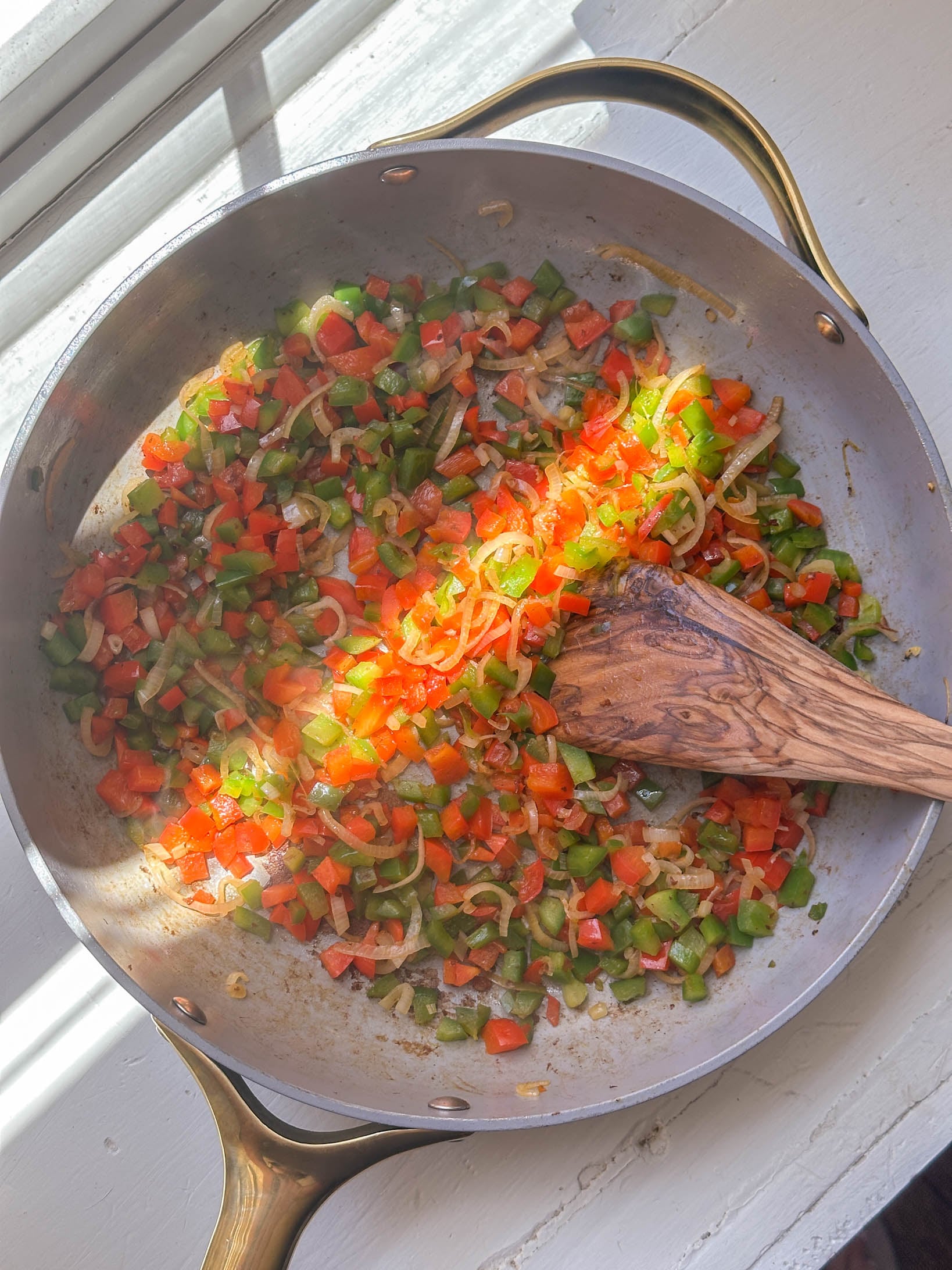 Diced red and green bell peppers with onions sautéing in a pan, stirred with a wooden spoon by a sunny window.