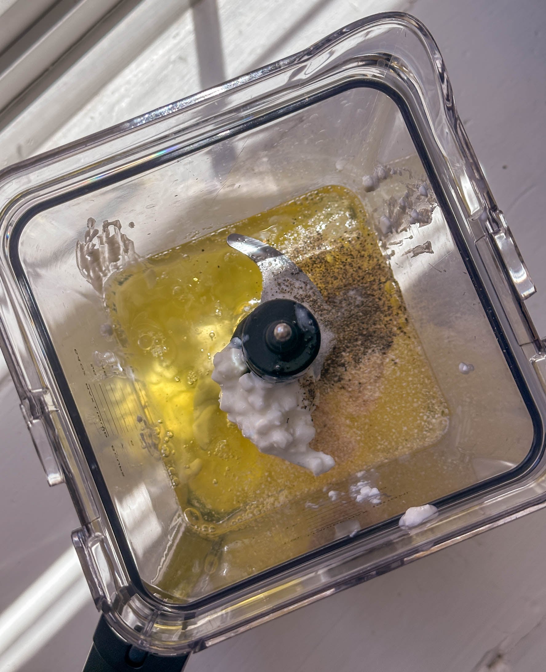 Top view of a blender with oil, seasonings, and a dollop of white ingredient inside on a sunny surface.