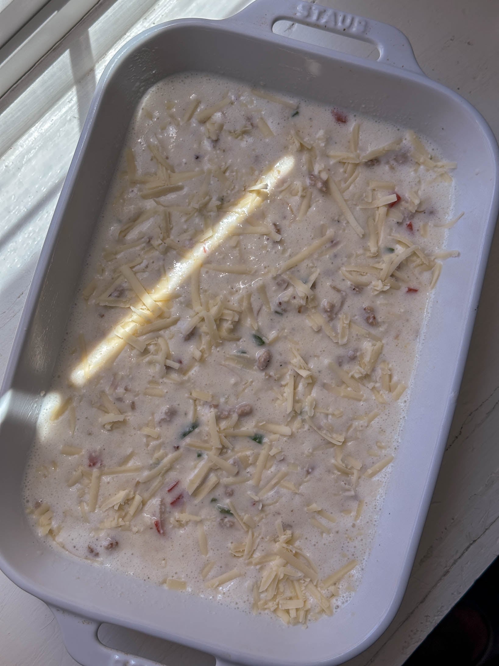 White casserole dish filled with an unbaked creamy mixture topped with shredded cheese, in sunlight.
