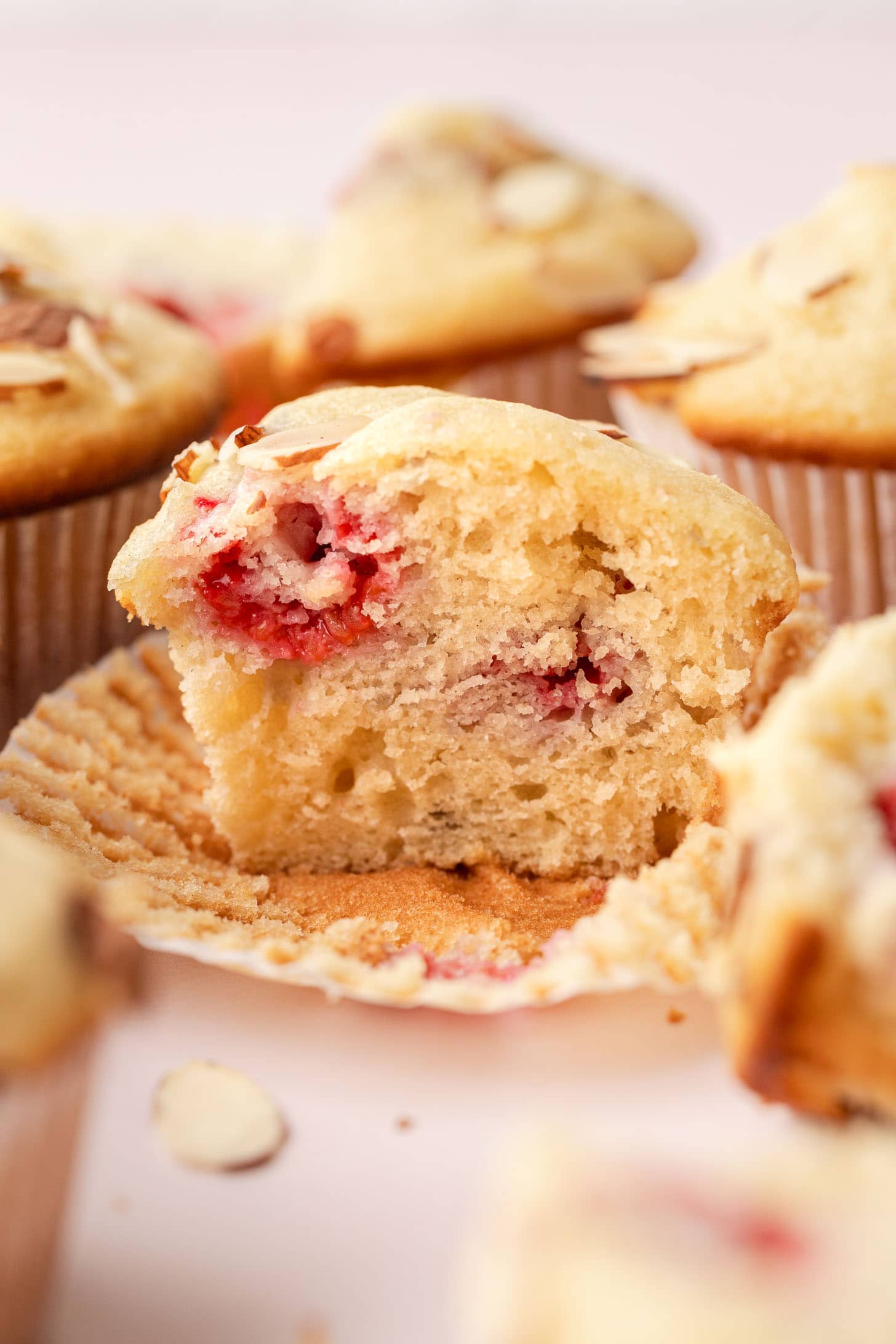 A close-up of a sliced muffin with visible raspberries and almond slices, surrounded by more muffins.