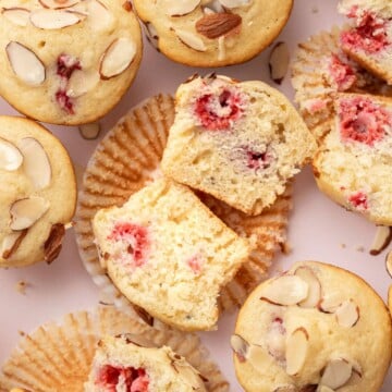Almond muffins with raspberry centers, some sliced open, on a light background with scattered almond slices.