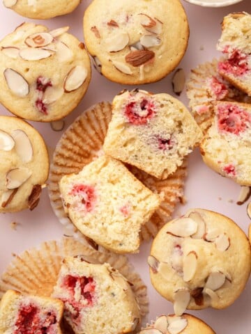 Almond muffins with raspberry centers, some sliced open, on a light background with scattered almond slices.