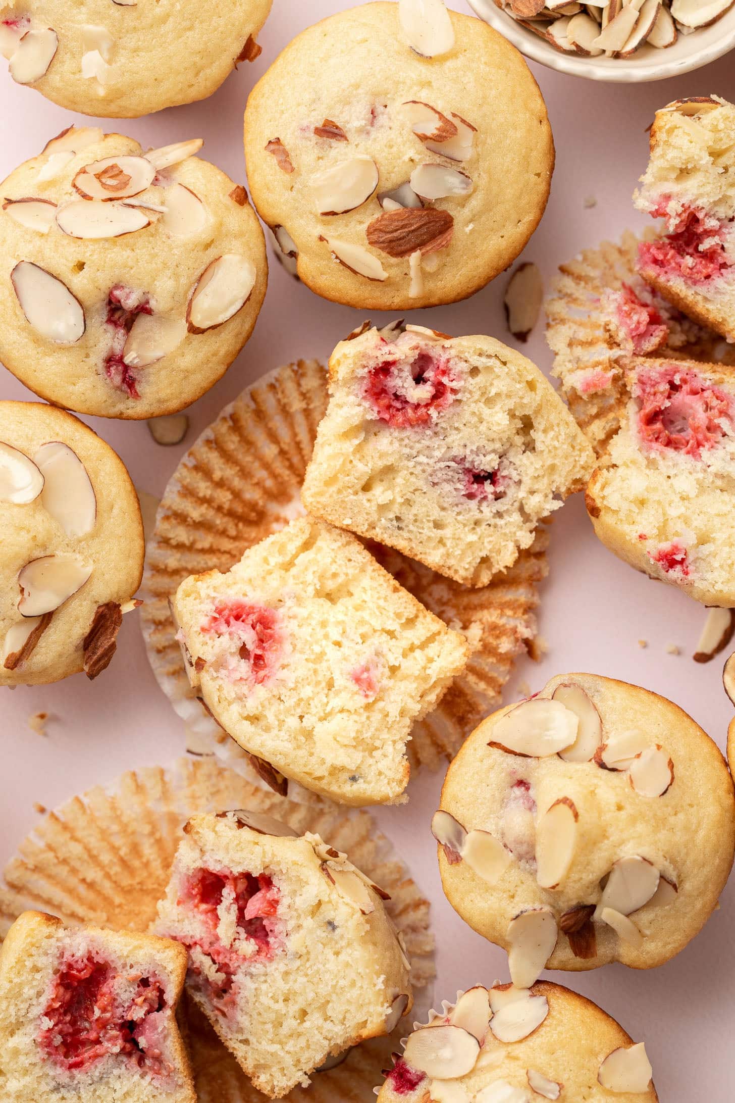 Almond muffins with raspberry centers, some sliced open, on a light background with scattered almond slices.