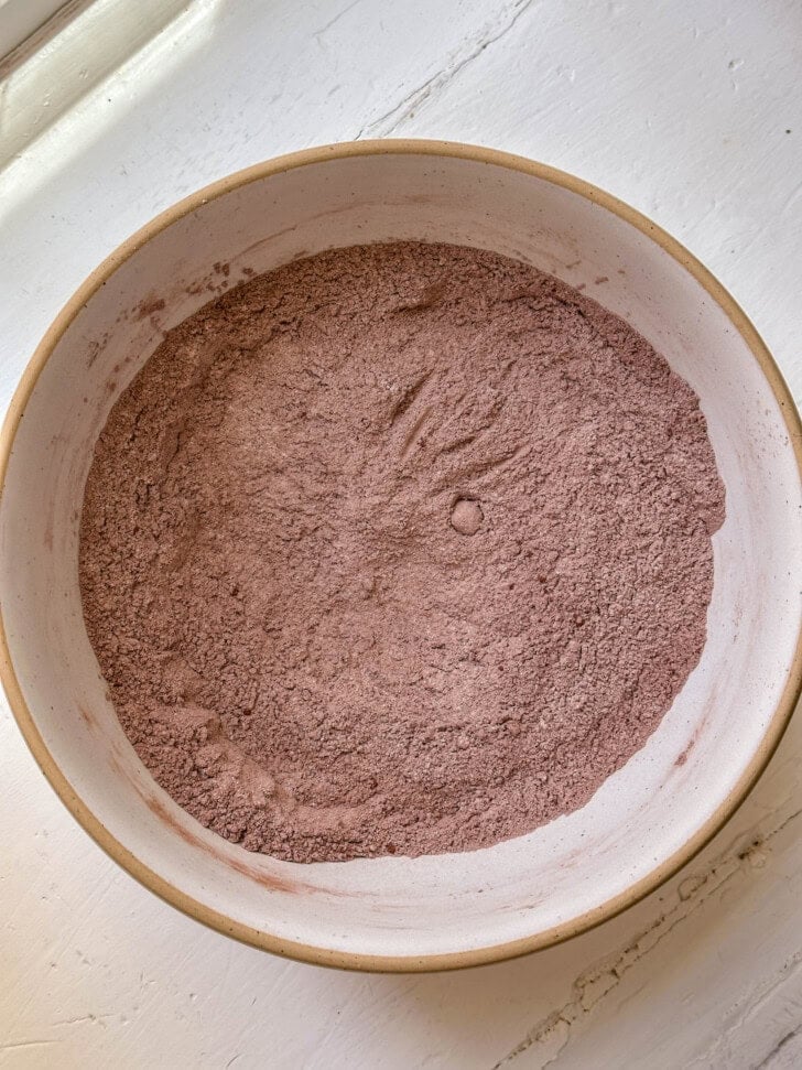 A white bowl filled with cocoa powder mix sits on a white surface near a window.
