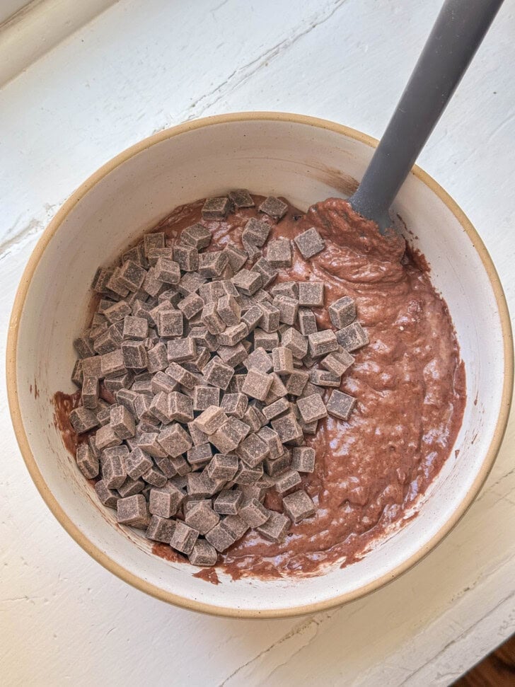 A mixing bowl with chocolate batter and chocolate chunks, being stirred with a gray spatula.