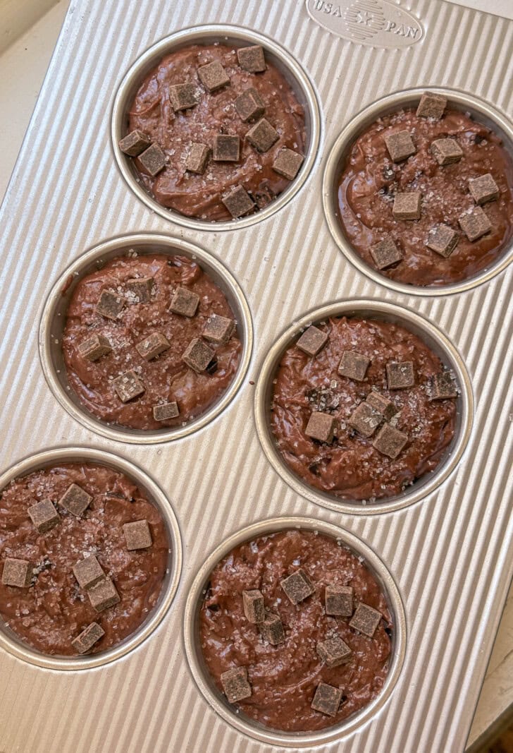 Six muffin cups filled with chocolate batter, topped with chocolate chunks and a sprinkle of salt.