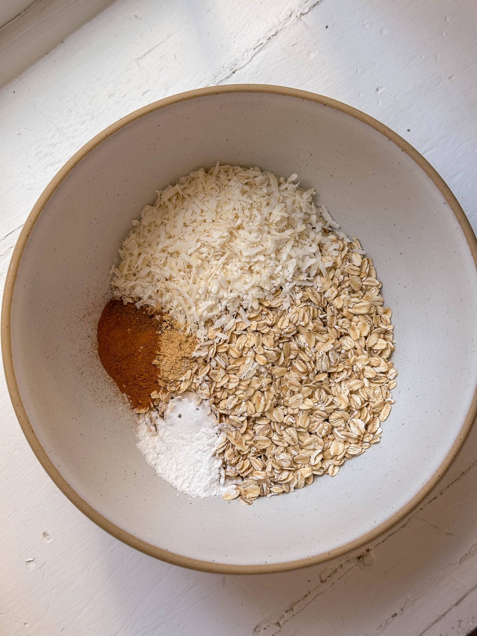 A bowl with oats, shredded coconut, cinnamon, and baking powder on a white surface.