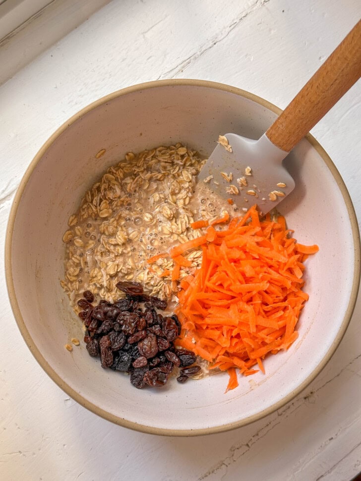 A bowl with oats, shredded carrots, raisins, and a spatula, ready to be mixed on a white surface.