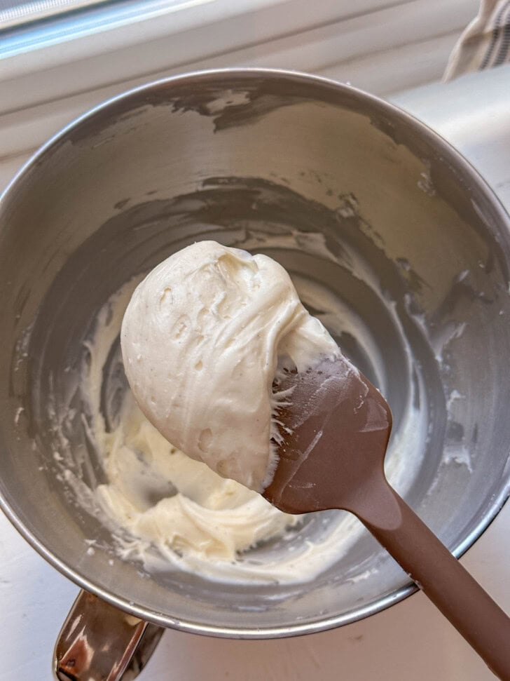 A spatula with creamy white frosting rests inside a metal mixing bowl on a white surface.