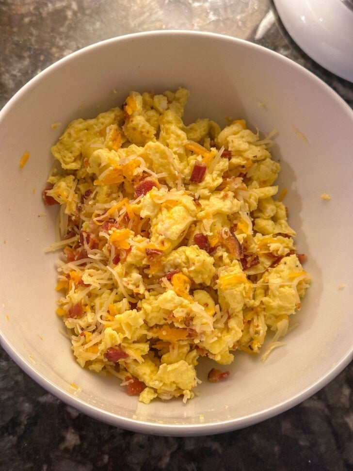 Scrambled eggs mixed with shredded cheese and bacon bits in a white bowl on a marble countertop.