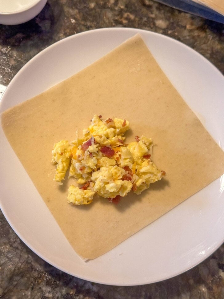 Scrambled eggs and bacon on a square sheet of dough, placed on a white plate.