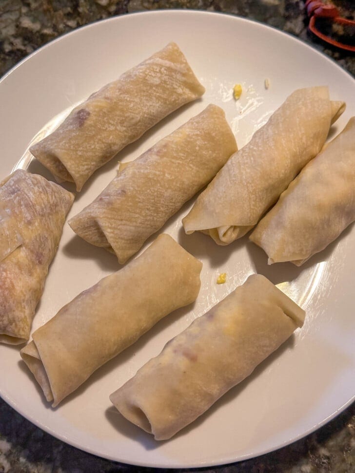Seven uncooked egg rolls are arranged on a white plate, ready to be cooked.