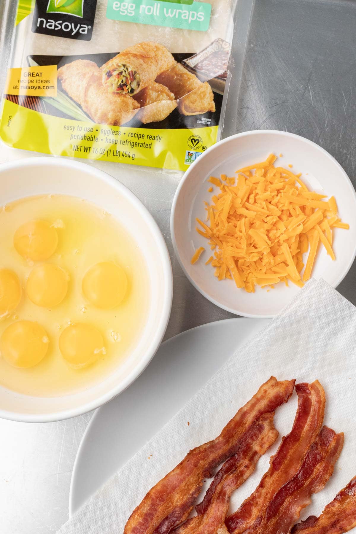 Bowl of eggs, shredded cheddar, cooked bacon, and a package of Nasoya egg roll wraps on a metal surface.