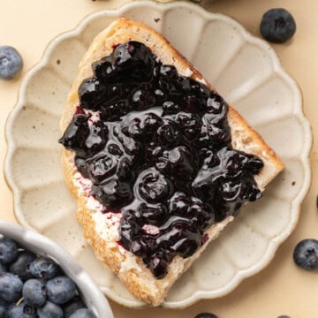 A slice of bread with blueberry jam on a plate, surrounded by fresh blueberries and a bowl of jam.