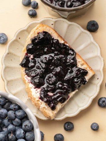 A slice of bread with blueberry jam on a plate, surrounded by fresh blueberries and a bowl of jam.