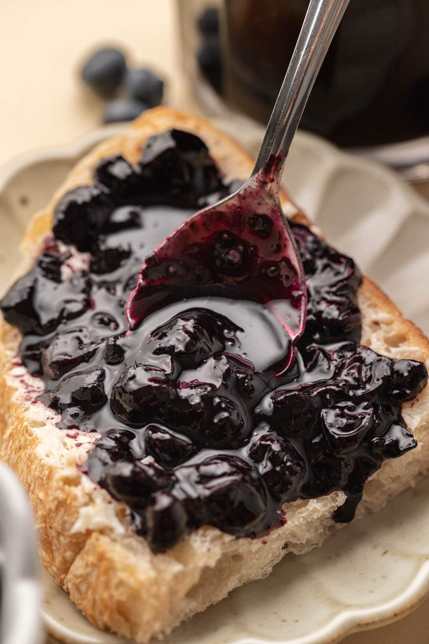 A spoon spreads thick blueberry jam on a slice of bread, with more blueberries blurred in the background.
