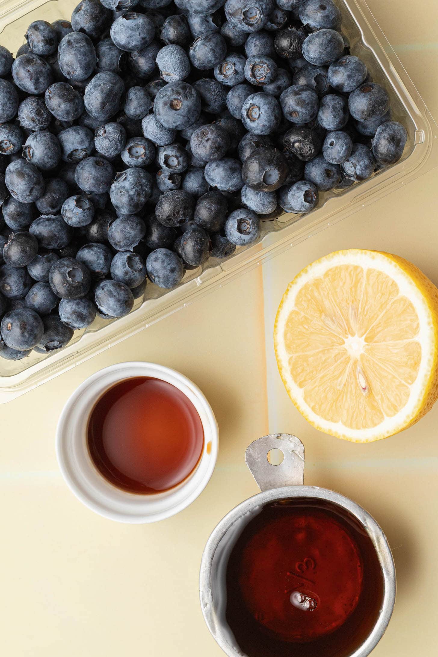 Fresh blueberries, a lemon half, and small containers of maple syrup and vanilla on a light surface.