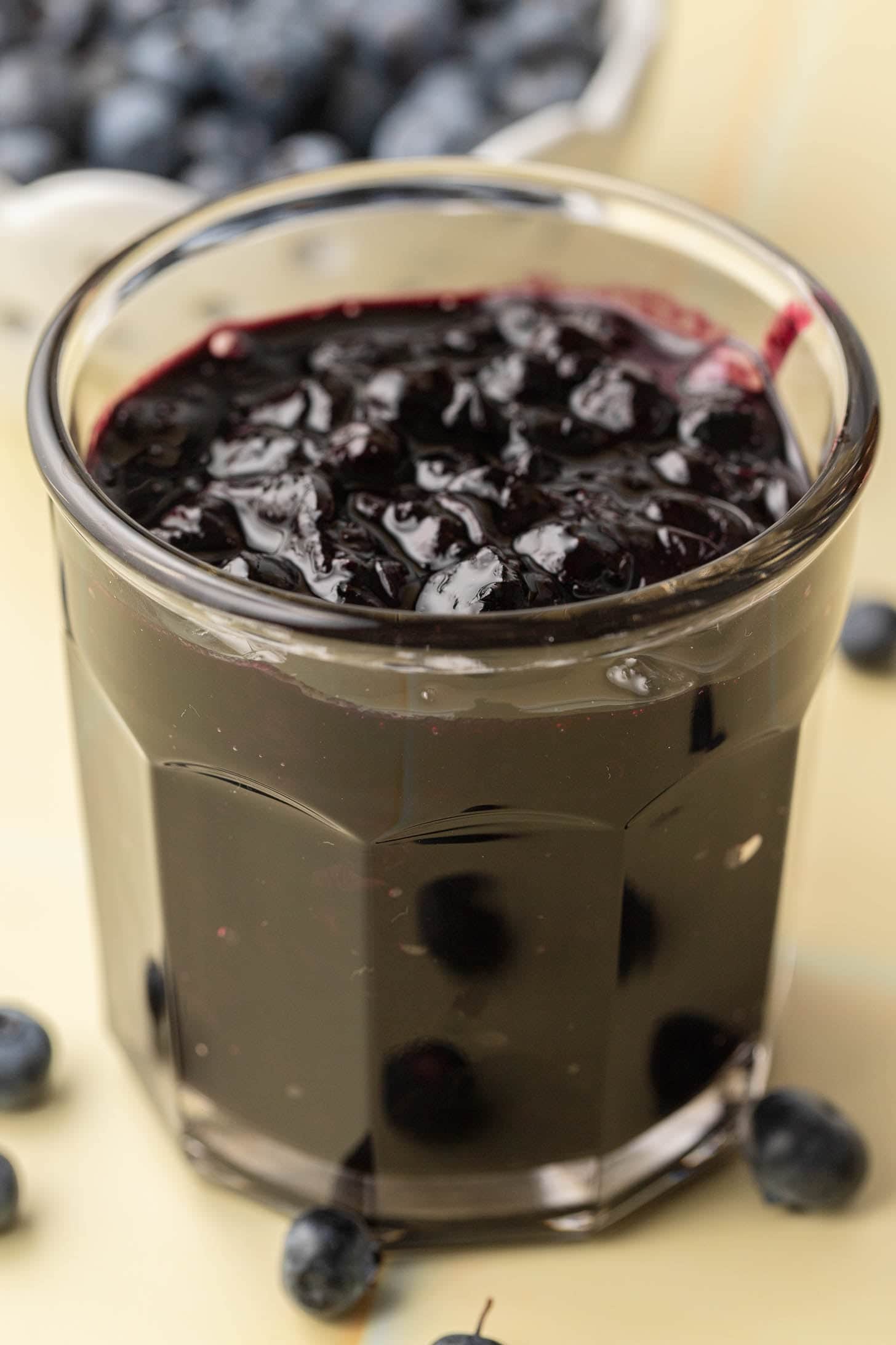 Close-up of a glass filled with blueberry jam, with fresh blueberries scattered around the base.