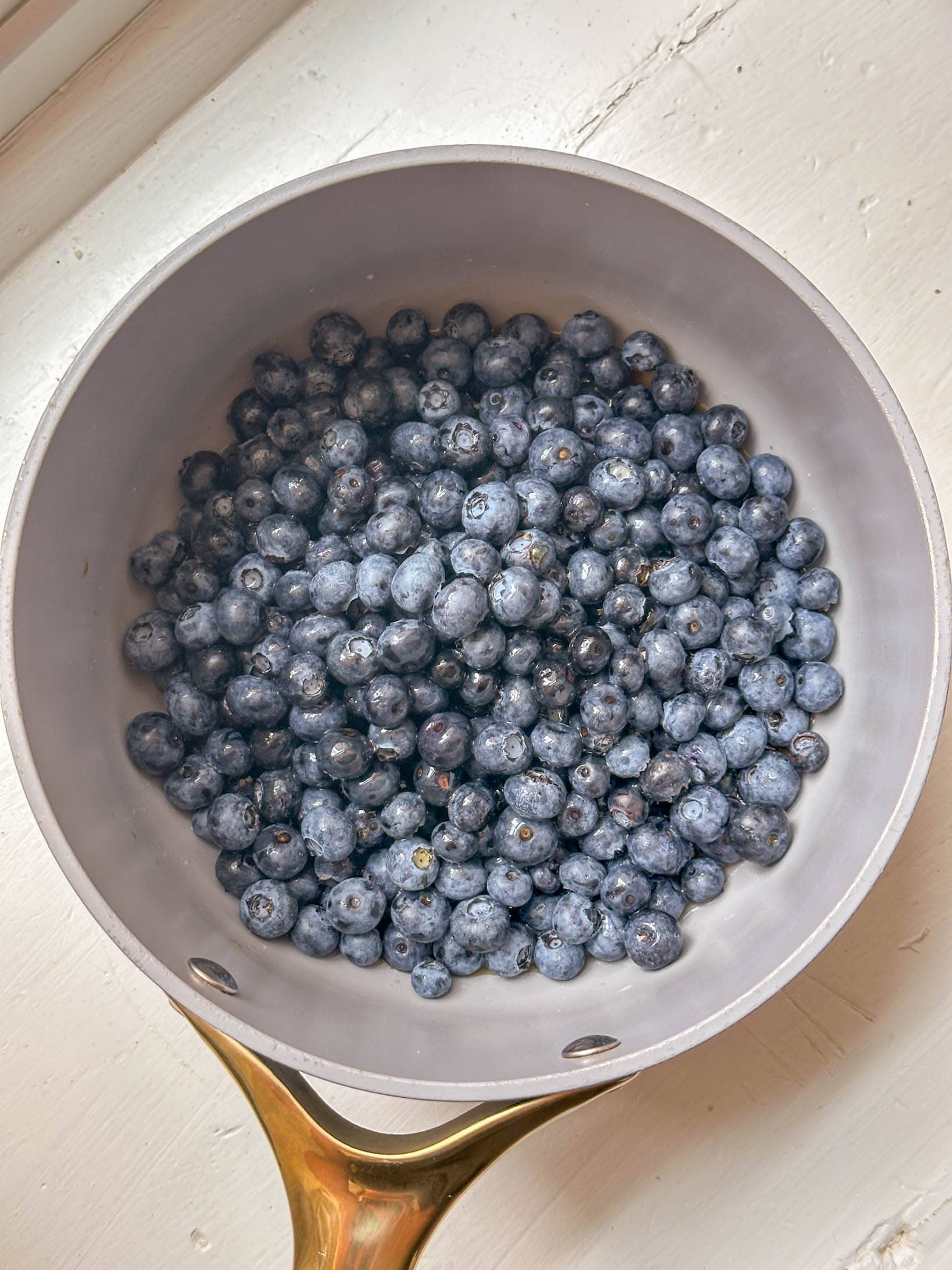 A saucepan filled with fresh blueberries sits on a light-colored surface near a window.