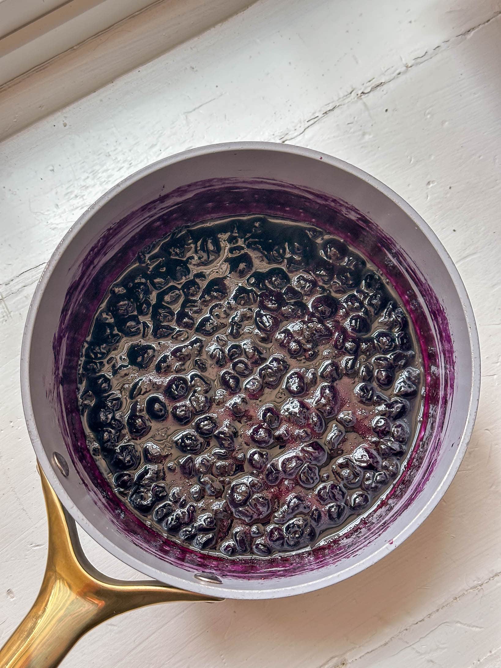 A saucepan with cooked blueberries sits on a white surface near a window.